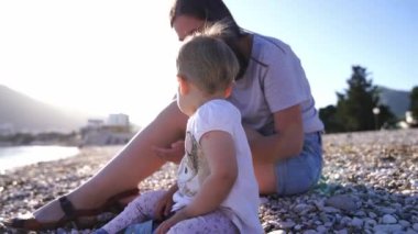 Küçük çocuklu bir anne kumsalda oturur ve denize çakıl taşları atar. Yüksek kalite 4k görüntü