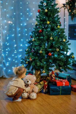 Oyuncak ayılı küçük bir kız altında hediyelerle bir Noel ağacının yanında çömeliyor. Yüksek kalite fotoğraf