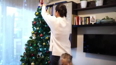 Küçük kız annesinin yanında yürüyor ve Noel ağacını oyuncaklarla süslüyor. Yüksek kalite 4k görüntü