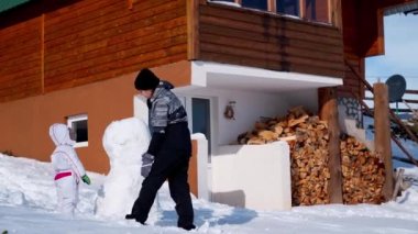 Anne ve küçük bir kız yakacak odunları olan ahşap bir evin yanında kardan adam yapıyorlar. Yüksek kalite 4k görüntü