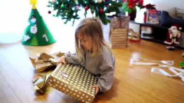 Noel ağacının yanındaki küçük kız hediye paketini açıyor, ambalaj kağıdını yırtıyor. Yüksek kalite 4k görüntü