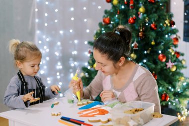 Küçük kız annesinin Noel kurabiyelerine renkli krema sürmesine yardım ediyor. Yüksek kalite fotoğraf