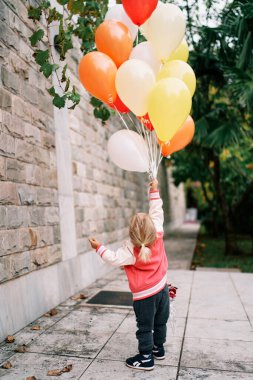 Elinde bir sürü balon olan küçük bir kız bahçedeki duvara yaslanmış ve kafasını kaldırmış. Arkadan bak. Yüksek kalite fotoğraf