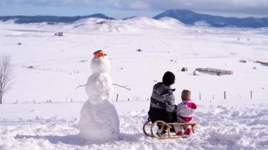 Anne, küçük bir çocuğa dağları gösteriyor. Kardan adamın yanında kızağın üstünde oturuyor. Yüksek kalite 4k görüntü