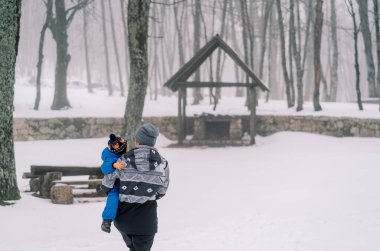 Anne, karla kaplı bir ormanda kucağında küçük bir çocuğu bir banka taşıyor. Arkadan bak. Yüksek kalite fotoğraf