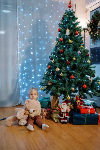 Oyuncak ayılı küçük kız Noel ağacının altında hediyelerin yanında yerde oturuyor. Yüksek kalite fotoğraf