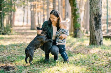Gülümseyen küçük kız annesine sarılıyor çömelmiş ve ormanda köpeğini okşuyor. Yüksek kalite fotoğraf