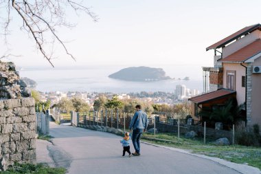 Baba ve küçük kızı, kasabanın dağ yolundan denizin arka planına doğru gidiyorlar. Arkadan bak. Yüksek kalite fotoğraf