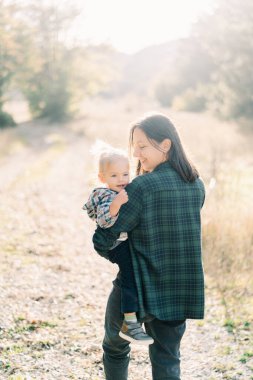Gülümseyen anne çimenlerde yürürken küçük kızını kollarında taşır ve ona bakar. Arkadan bak. Yüksek kalite fotoğraf