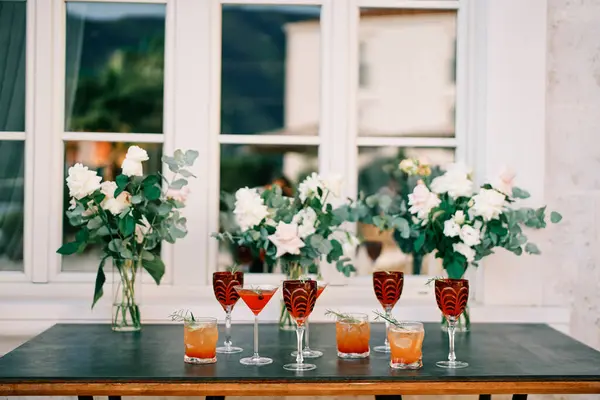 Glasses with alcoholic drinks stand on the table near bouquets of flowers in vases near the window of the building. High quality photo