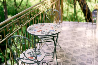 Çiçekli süslemeleri olan yuvarlak bir masa ile dövülmüş sandalyeler bahçedeki fayanslı terasta duruyor. Yüksek kalite fotoğraf