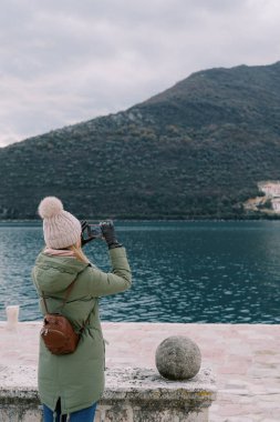 Kız deniz kıyısındaki taş çitin yanında duruyor ve akıllı telefonuyla dağların fotoğraflarını çekiyor. Arkadan bak. Yüksek kalite fotoğraf