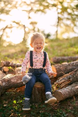 Küçük kız elinde oyuncakla ormanda bir kütüğe oturur ve gözleri kapalı güler. Yüksek kalite fotoğraf