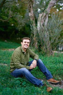 Parkta yeşil çimlerin üzerinde oturan gülümseyen genç adam. Yüksek kalite fotoğraf