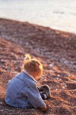 Küçük kız deniz kıyısında oturur ve küçük çakıl taşlarını kazar. Yan görüş. Yüksek kalite fotoğraf