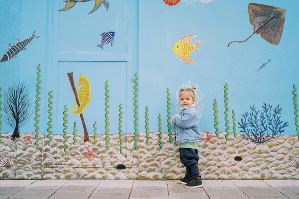 Маленькая девочка стоит, прислонившись к стене с красочными рисунками. Высокое качество фото