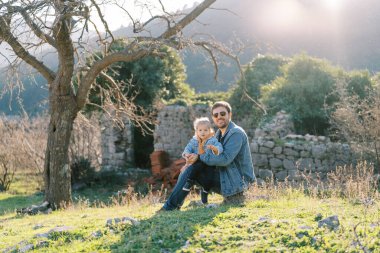 Babam kollarında küçük bir kızla koruluktaki taş bir çitin yanında oturuyor. Yüksek kalite fotoğraf
