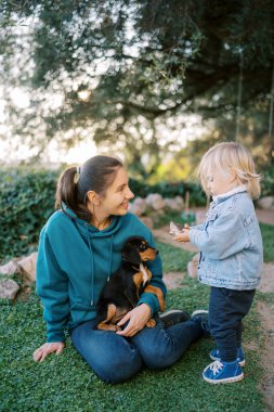 Küçük kız, gülümseyen bir annenin yanında dizlerinin üzerinde bir köpek yavrusuyla çimlerde oturuyor. Yüksek kalite fotoğraf