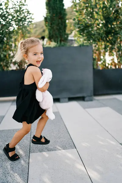 Küçük gülen kız bahçedeki fayansları koşarak geçiyor, oyuncak tavşana sarılıyor. Yüksek kalite fotoğraf