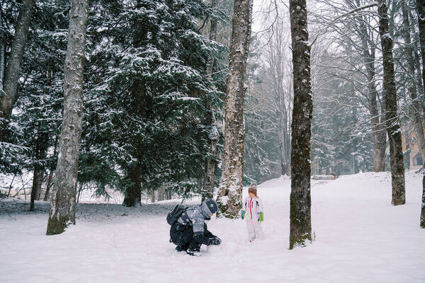 Little girl stands in a snowy forest and looks at her mom making a snowman. High quality photo