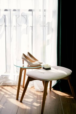 Gelin ayakkabıları, içinde alyanslar olan bir kutunun yanında duruyor. Odadaki rahat bir sandalyenin yanında cam bir masanın üzerinde. Yüksek kalite fotoğraf