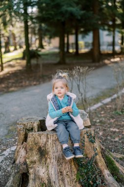 Küçük kız parktaki sarmaşıkla kaplı bir kütüğe oturur. Yüksek kalite fotoğraf