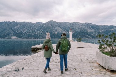 Sırt çantalı gezginler rıhtımda el ele tutuşarak deniz fenerine yürüyorlar. Arkadan bak. Yüksek kalite fotoğraf