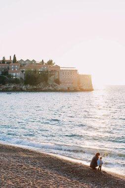 Annem ve küçük bir kız sahilde oturmuş Sveti Stefan adasının yanındaki denize bakıyorlar. Karadağ. Yüksek kalite fotoğraf