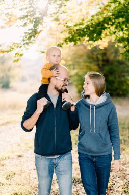 Omuzlarında küçük bir oğlu olan anne ve baba parkta yürüyüp birbirlerine bakıyorlar. Yüksek kalite fotoğraf