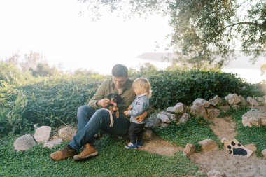 Küçük kız babasının yanında duruyor çimenlikte oturan bir köpek yavrusuyla kucağında oyuncak kokluyor. Yüksek kalite fotoğraf