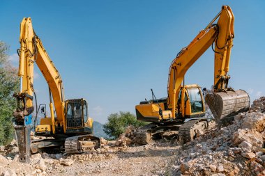 Bir çift güçlü sarı kazıcı kayalık bir inşaat sahasının ortasında duruyor. Mekanik kollar hazır, makinelerin ağır kaldırma ve toprak hareket kabiliyetini vurguluyorlar.
