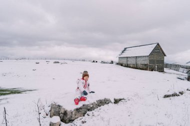 Bir tepe çiftliğinin yakınındaki karlı bir çayırda oturan küçük kız. Yüksek kalite fotoğraf