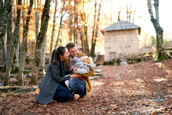 Laughing mom tickles little girl on dad lap squatting in autumn park. High quality photo