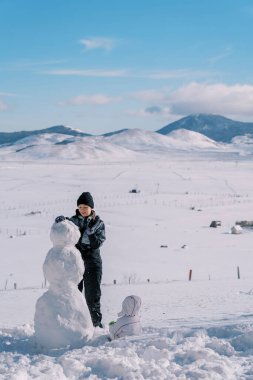 Küçük çocuk karda oturur ve annesinin dağ vadisinde bir kardan adam yapmasını seyreder. Arkadan bak. Yüksek kalite fotoğraf