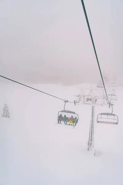 Tourists in colorful ski suits ride uphill through the fog on a chairlift above a snowy forest. High quality photo
