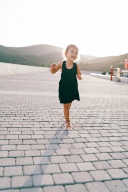 Küçük gülen kız fayanslı bir yolda çıplak ayakla yürüyor, dans ediyor ve kollarını sallıyor. Yüksek kalite fotoğraf