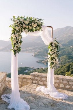 Beyaz güllerle süslenmiş düğün kemeri denizin üstünde bir gözlem güvertesinde ve dağlık bir sahilde duruyor. Yüksek kalite fotoğraf