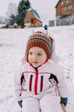 Küçük gülümseyen kız karlı bir tepenin dibindeki kızağa oturur ve yana bakar. Yüksek kalite fotoğraf