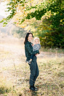 Gülümseyen anne, kollarında küçük bir kızla bir ağacın yanında yarı dönüşmüş duruyor. Yüksek kalite fotoğraf