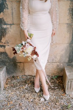 Elinde buket olan gelin, antik bir binanın taş duvarına yaslanmış duruyor. Kırpılmış. Yüzsüz. Yüksek kalite fotoğraf