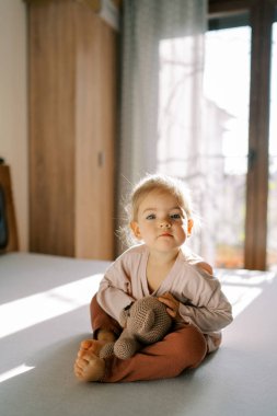Küçük bir kız yatak odasında yatakta oturmuş, oyuncak ayıya sarılıyor ve öne eğiliyor. Yüksek kalite fotoğraf