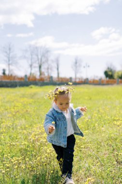 Çelenk takmış küçük bir kız çiçekli bir çimenlikte yürüyor, ayaklarına bakıyor. Yüksek kalite fotoğraf