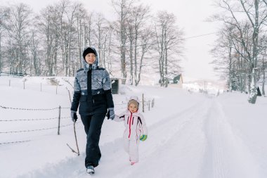 Anne ve küçük bir kız, ormanın kenarında karlı bir yolda el ele yürüyorlar. Yüksek kalite fotoğraf