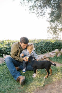 Kollarında küçük bir kızla gülümseyen baba çimlere oturur ve topu yavru köpeğe uzatır. Yüksek kalite fotoğraf