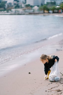 Eldivenli küçük kız deniz kenarındaki kumlu bir sahilden çöp topluyor. Yüksek kalite fotoğraf