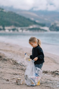 Küçük kız deniz kenarındaki kumlu bir sahilde plastik şişeleri bir çöp torbasına koyuyor. Yüksek kalite fotoğraf