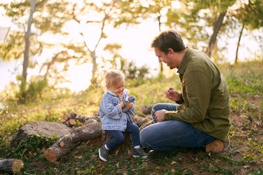 Küçük kızıyla gülen baba ormandaki güneşli bir çayırda kütüklerin üzerinde oturuyor. Yüksek kalite fotoğraf