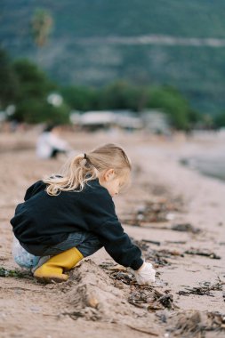 Küçük kız kumsalda çantasıyla çömelirken kumdan çöp topluyor. Yüksek kalite fotoğraf