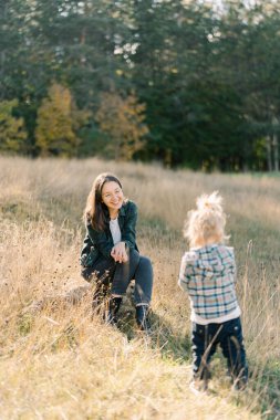 Gülümseyen anne bir taşın üzerine oturur ve açıklıkta duran küçük kızına bakar. Yüksek kalite fotoğraf