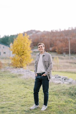 Elleri pantolonunun cebinde gülümseyen genç adam açıklıktaki sarı bir ağacın yanında duruyor. Yüksek kalite fotoğraf